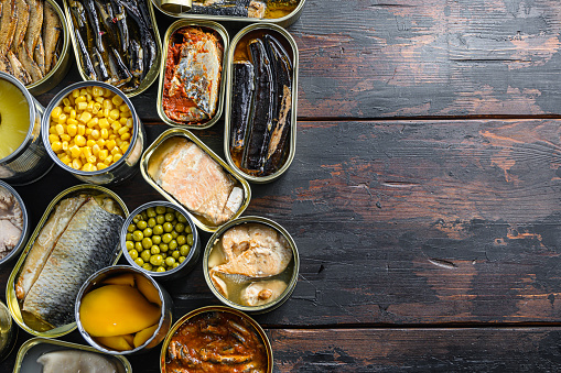 Canned preserves  food in tin  open cans. with fresh organic ingridients Saury, mackerel, sprats, sardines, pilchard, squid, tuna pinapple, corn, peas, mango ,  beans,   over  old wood dark textured background  top view space for text.