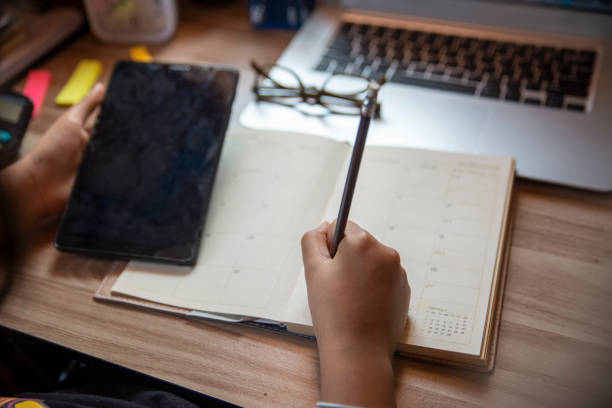 arbeiten von zu hause aus konzept. planer mit tablet und schreiben, um die tägliche agenda im kalenderbuch zu planen. hand der frau markieren und notierte zeitplan (urlaubsreise) auf tagebuch am home-office-schreibtisch. kaffeeplatz auf dem tisch - working at home calendar laptop women stock-fotos und bilder