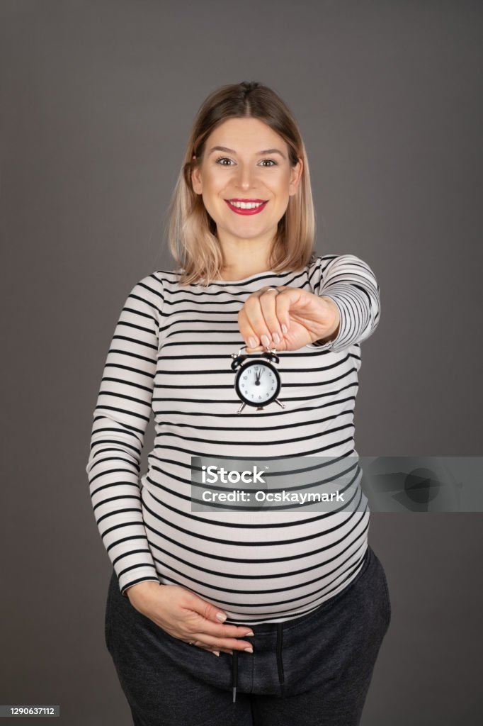 Pregnant woman holding a clock Pregnant woman in third trimester waiting for childbirth, holding a clock on grey background Abdomen Stock Photo