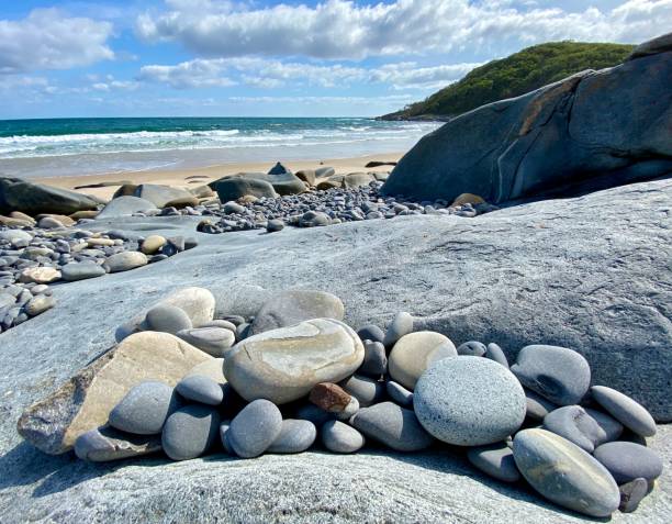 Horizontal landscape photo of worn, smooth, rounded grey granite boulders and rock platforms on the beach at Granite Bay. A forested headland and the blue Pacific Ocean can be seen in the background under a cloudy blue sky on a sunny day in Summer