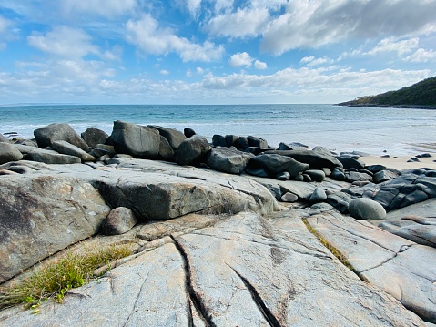 Horizontal landscape photo of the Pacific Ocean, a large smooth grey granite rock platform, worn granite boulders and part of a forested headland under a cloudy blue sky on a sunny day in Summer at Granite Bay in the Noosa Heads National Park