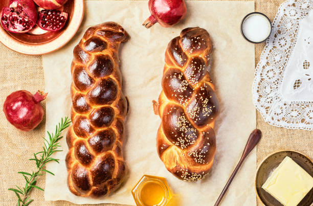 Challah bread, freshly baked sweet braided bread on parchment, sackcloth background. Pomegranates, apples, butter and honey for bread on the table. Top view, product layout, hanukkah celebration stock photo