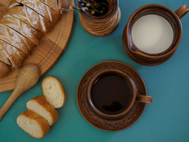 Flat lay of beautifully served breakfast in handmade earthenware against a green background. Fresh delicious pastries with bird cherry filling, a cup of hot aromatic tea, a crispy French baguette, milk in an earthen jug complemented by willow in a vase and a wooden spoon. View from above