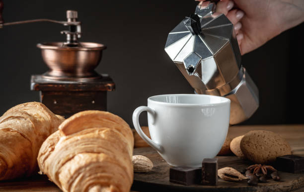istock A hand is pouring black aromatic coffee from a geyser coffee maker into a white cup. Fresh croissants and a coffee grinder are next to the mug on the wooden table 1290653140