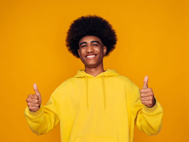 joven sonriente con sudadera amarilla con capucha y con los pulgares hacia arriba - afro man fotografías e imágenes de stock