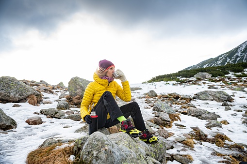 A woman traveler in yellow down jacket is resting with a metal mug of hot tea on a cold windy day.