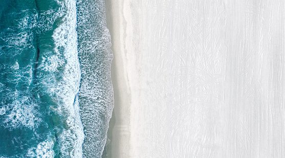 Drone view of ocean blue water with sea waves rolling on an a large and empty white beach. Copy space on the right side.