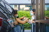 Asian man in protective mask taking food bag and coffee with woman waitress wearing face mask and face shield at drive thru during coronavirus outbreak.