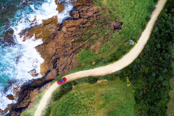 Driving on a seaside road approaching a beach, seen from above Red car moving on a winding coastal road approaching a beautiful beach with sea waves and blue water. Green forest and rocks for a perfect summer destination. Aerial view. drone point of view road stock pictures, royalty-free photos & images