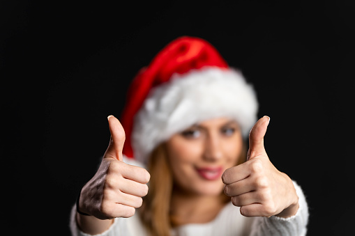 Beautiful young woman wearing Santa hat looking at camera and showing thumbs up hand sign on black background. The female is of caucasian ethnicity, wearing santa claus christmas hat and is dressed in casual clothing. The scene is situated in a professional photo studio and is set in front of a black background / backdrop. The picture is taken with full frame Sony A7III camera.