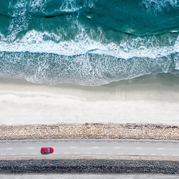Red car moving on a straight coastal road approaching a beautiful beach with white sands and sea waves. Perfect destination for a summer escape to the sea.