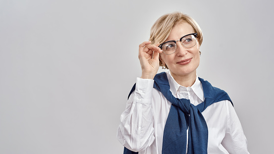 Portrait of elegant middle aged caucasian woman wearing business attire adjusting her glasses, smiling aside while standing isolated over grey background. Front view. Web Banner