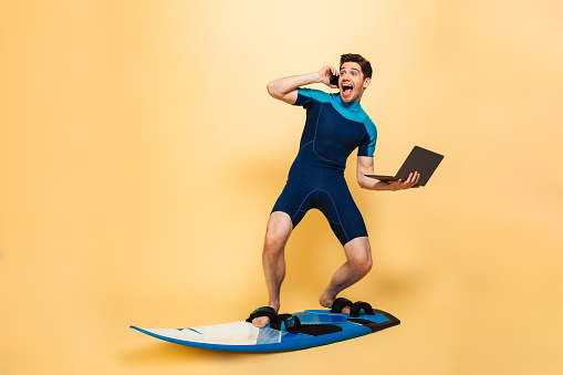 Photo of a excited young man dressed in swimsuit isolated over yellow background on surfing board talking by phone using laptop computer.