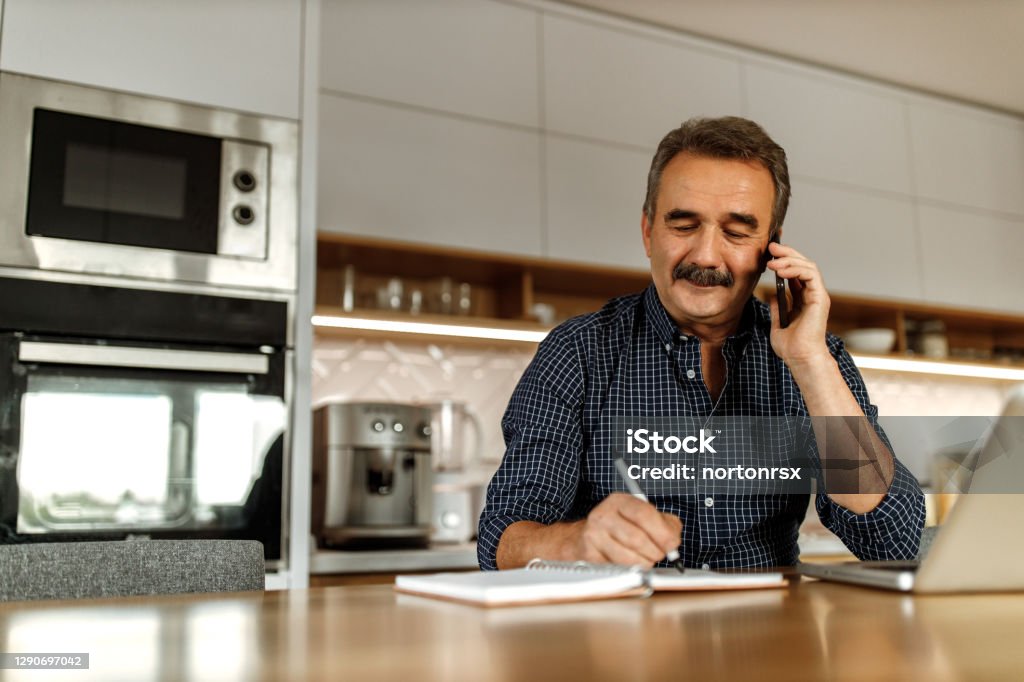 Man talking with someone over the phone. Adult man making phone call, while reading and writing some words in his notebook. Using Phone Stock Photo