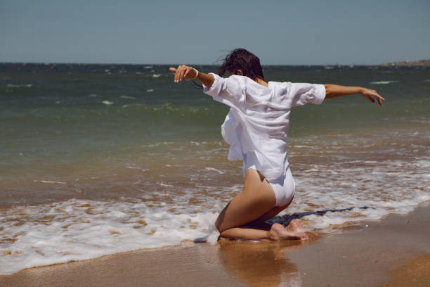 woman in a white bathing suit and a straw hat stands on an empty sandy beach