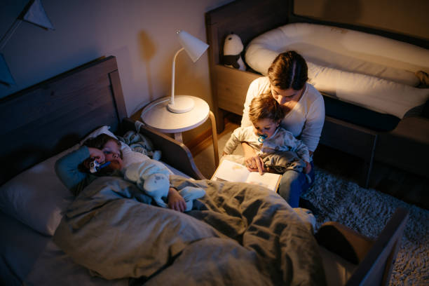 Mother reading a book to her two sons Young mother is reading a book to her two sons for a good night sleep. Night time with reading lamp. Horizontal photo. bedtime stock pictures, royalty-free photos & images