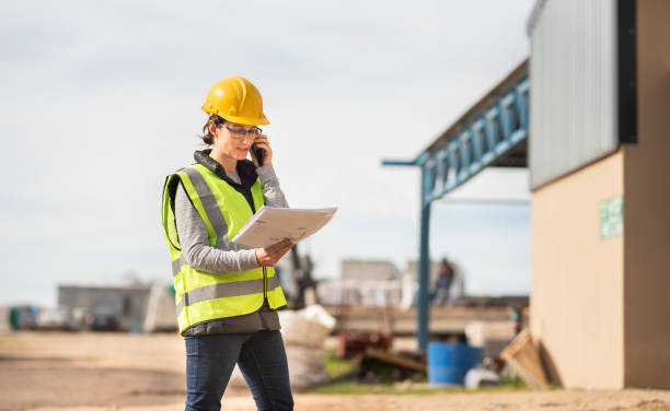 ingénieur féminin parlant au téléphone et affichant des plans sur un chantier - architect construction engineer standing photos et images de collection