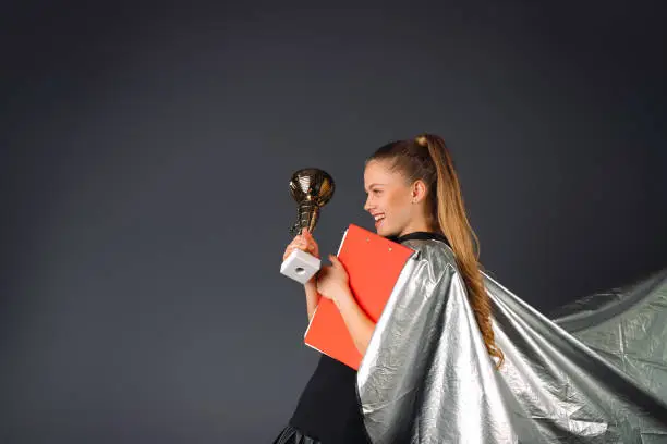 Superheroine, businesswoman with a red folder and a champions cup in hands being satisfied of trophy on a gray background. High quality photo
