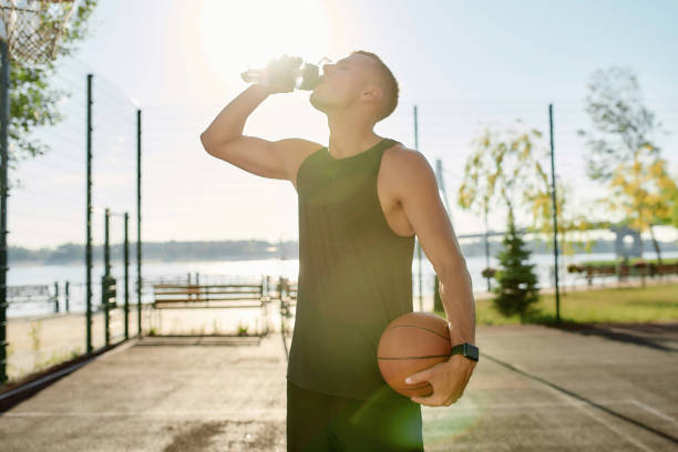 müde sportliche junge kerl trinkwasser ausform flasche, halten basketball, während posiert auf dem platz an einem sonnigen tag - basketball teenager nature outdoors stock-fotos und bilder