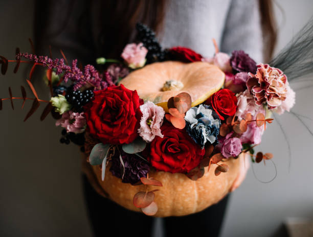 Nice young woman holding orange carved pumpkin filled with blossoming roses, carnations, eucalyptus flowers in passionate red colors, halloween themed design stock photo