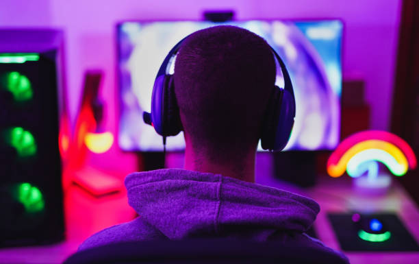 joven que lleva auriculares y jugar videojuegos de ordenador en línea - inicio aislado para coronavirus - videojuego fotografías e imágenes de stock
