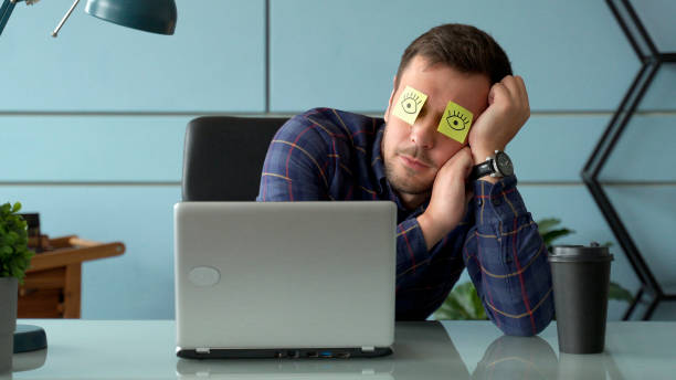 Tired comic male manager pretends working sleeping with stickers on face sits at desk with laptop Tired comic male manager pretends working sleeping with stickers on face sits at desk with laptop lazy construction laborer stock pictures, royalty-free photos & images