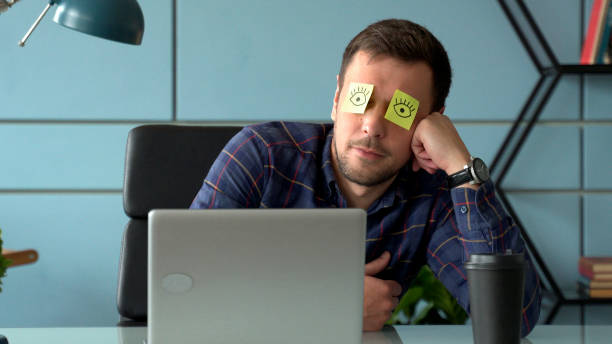 Tired comic male manager pretends working sleeping with stickers on face sits at desk with laptop Tired comic male manager pretends working sleeping with stickers on face sits at desk with laptop lazy construction laborer stock pictures, royalty-free photos & images