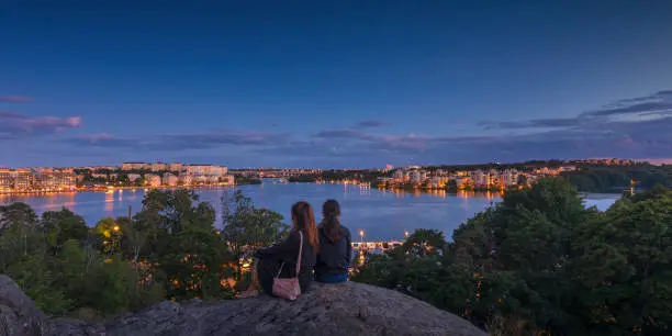 Two women sitting at a viewpoint and looking out over lake Mälaren towards Kungsholmen and Bromma in Stockholm on a summer evening.