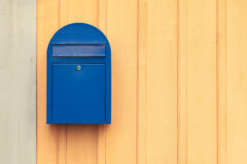 A classic designed blue mailbox in front of the house fence and flower garden (as blurred background). Close-up and selective focus.