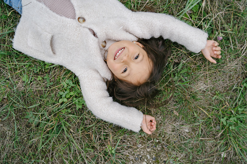 Little child in a cute white sweater. Girl spend time in a park. Kid luing on a grass.