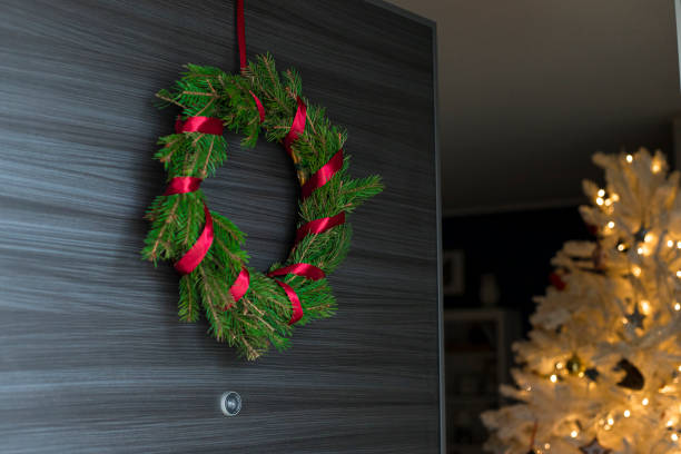 Foraged diy pine wreath with red ribbon on door opening with white, illuminated christmas tree inside the house.