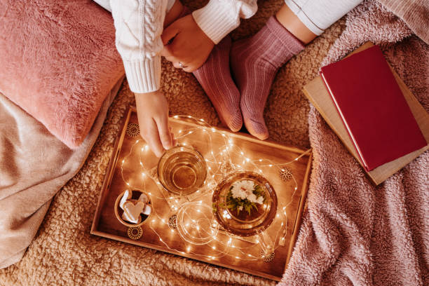 Unrecognizable girl in cozy woolen socks with woolen blankets and light chain drinking blooming flower tea sitting on the bed
