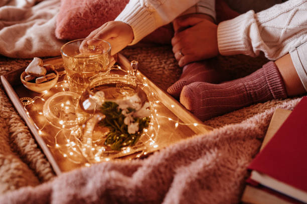 Unrecognizable girl in cozy pink woolen socks and pullover with winter blankets enjoying her blooming flower tea and light chain on the bed