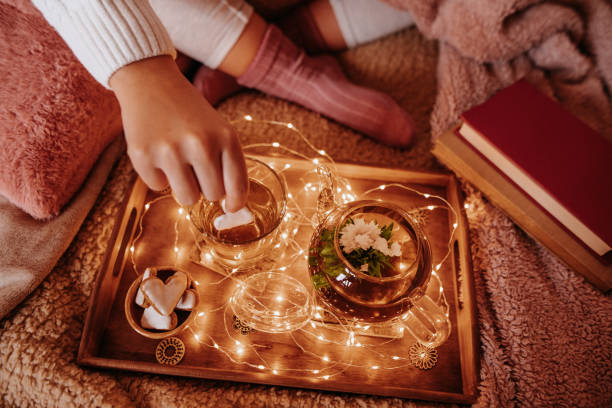 Unrecognizable young girl in cozy woolen socks with winter blankets enjoying her breakfast with blooming flower tea and light chain on the bed
