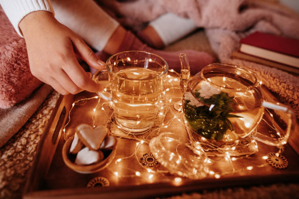 Unrecognizable girl in cozy woolen socks with winter blankets and light chain drinking blooming flower tea sitting on the bed