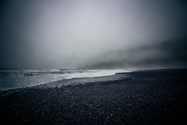 A misty, low angle shot shows waves creep into the black sand coast of a beach near Vik.