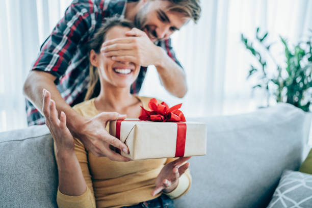 Cheerful young woman receiving a gift from her boyfriend. Handsome young man is standing behind his girlfriend with a gift in one hand and covered her eyes with the other hand. Focus is on the gift. gift for girlfriend stock pictures, royalty-free photos & images