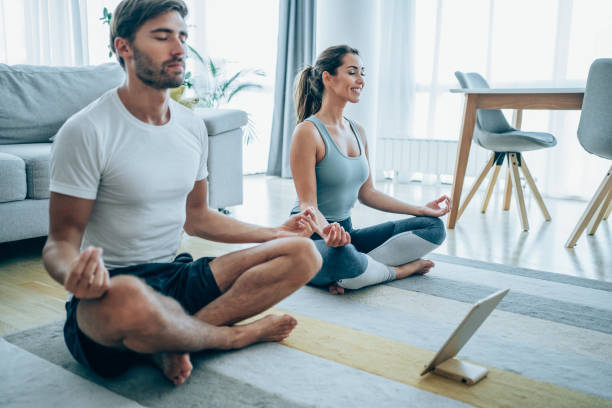 Young couple meditating in Lotus position at home. Beautiful young couple sitting on the floor and practicing yoga. Sporty couple using digital tablet for online yoga lessons. mindful happy home stock pictures, royalty-free photos & images