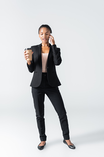 african american businesswoman with paper cup talking on smartphone isolated on grey