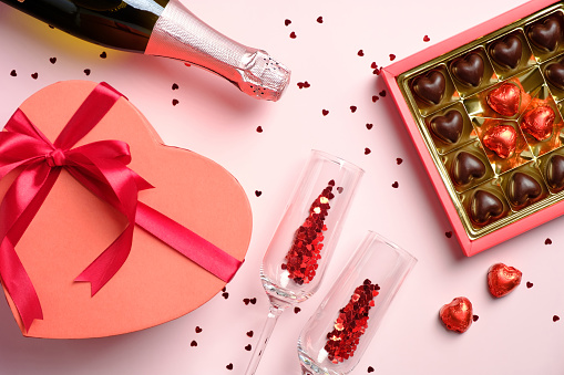 Happy Valentines day celebration concept. Flat lay composition with heart shaped box decorated red ribbon bow, sweets, wine and glasses on pink table. Top view.