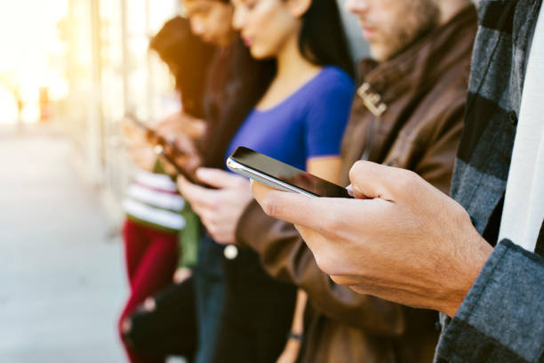 fila de jóvenes adultos en sus teléfonos inteligentes - diversidad - comunicación - dispositivos electrónicos - al aire libre - staring fotografías e imágenes de stock