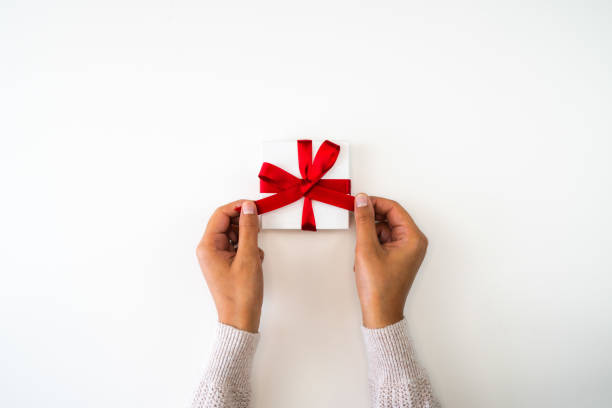 Holiday or Christmas background or backdrop image close up of hands from a mixed race African American woman wearing a sweater tying a red ribbon bow onto a small white gift box with copy space. stock photo