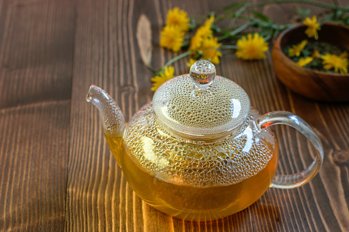 Glass teapot with golden dandelion tea on the wooden table.