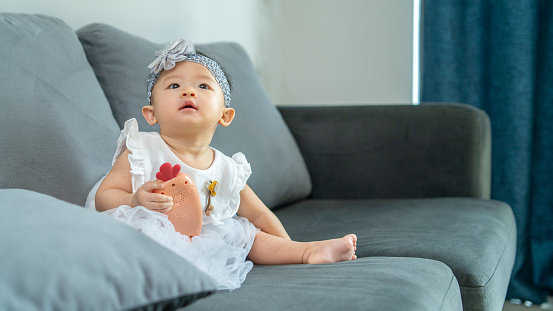Cutie baby girl holding toy looking away. She is sitting on the sofa in the living room.