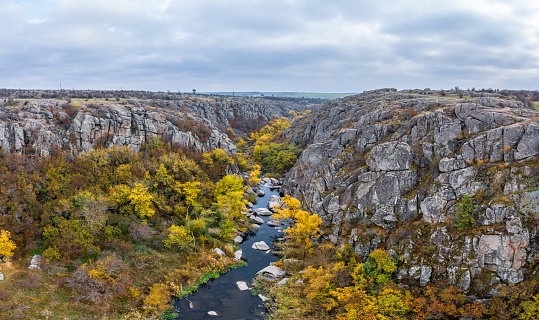 A stream flows in the Aktovsky Canyon, Ukraine. Autumn trees and large stone boulders around. Aerial panoramic drone shot