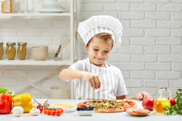 little boy in chef hat and an apron cooking pizza little boy in chef hat and an apron cooking pizza in the kitchen. child adding cheese into pizza pepper cake stock pictures, royalty-free photos & images