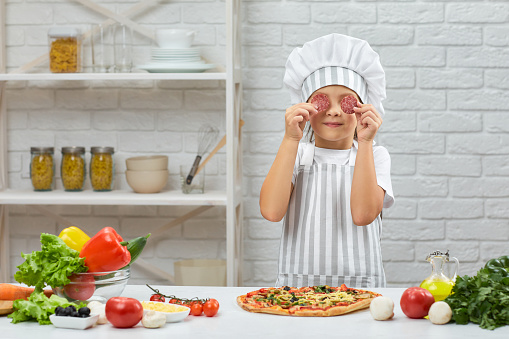 little girl in chef hat and an apron cooking pizza in the kitchen. the child holding sausage slices in front of his eyes. having fun