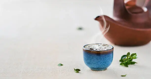 Cup of hot japanese green tea. Selective focus on the cup. Steam rises from the cup of tea.Teapot and green leaves on the table. Close-up, with copy space. Tea party idea, minimalism