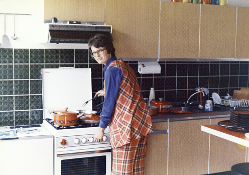 Vintage analog 1970's image of a mother wearing an orange squared pant and vest cooking dinner on a gas stove.