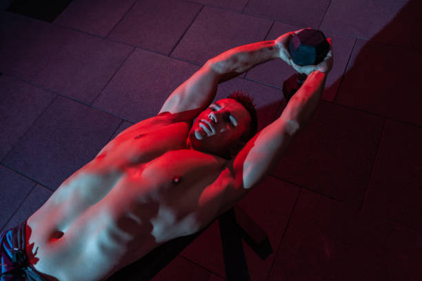 Muscular man exercise with dumbbell lieas on bench in red-blue gradient light in dark gym hall